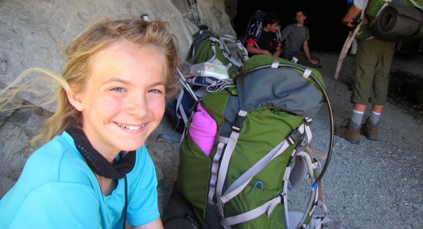 a young person sits beside their backpack and smiles at the camera